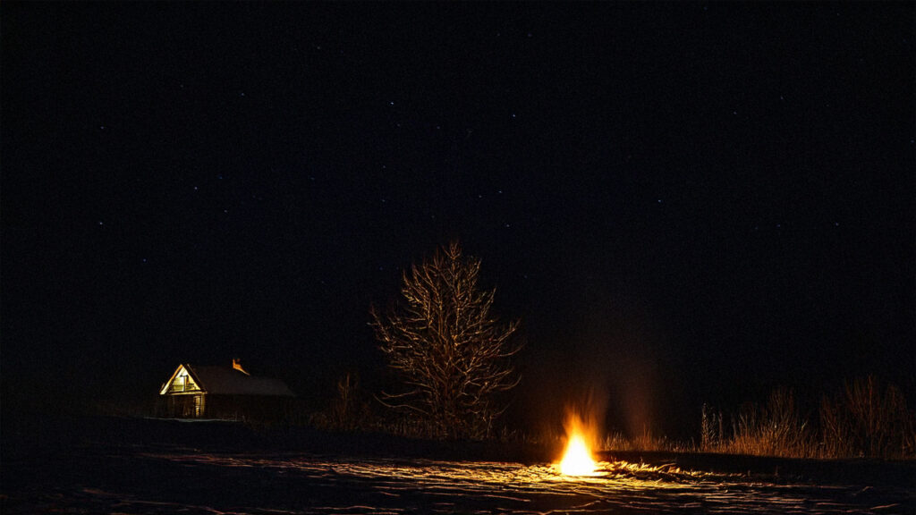 Photo of dark winter night with small fire in foreground and a house in the distance